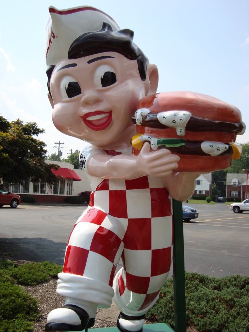A cheerful, cartoonish mascot in a checkered outfit holds a large hamburger, standing outside a restaurant.