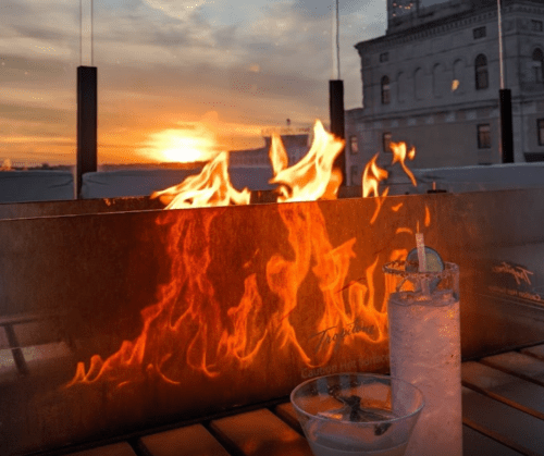 A sunset view from a rooftop bar, featuring flames and cocktails on a table.