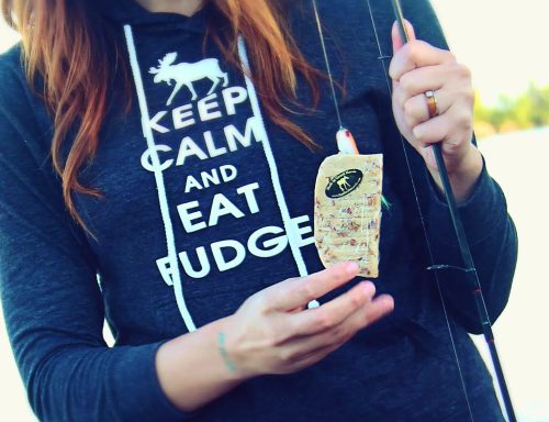 A person in a hoodie holds a fishing lure by a lake, promoting a lighthearted fishing theme.