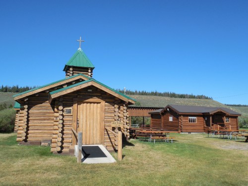 A rustic log cabin church with a green roof, next to a larger log building, set in a grassy area under a clear blue sky.