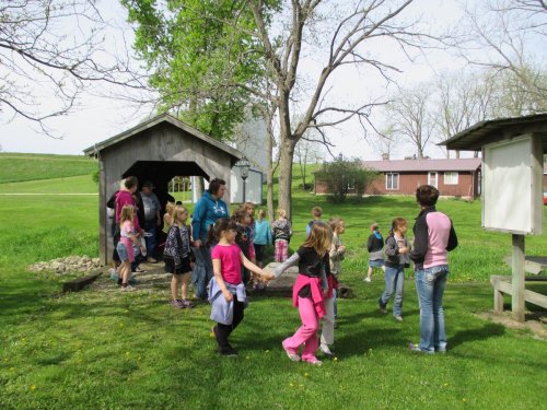 A group of children and adults gather near a wooden structure in a grassy area, with trees and a building in the background.