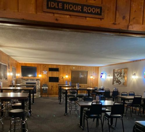Interior of a dimly lit bar room with wooden walls, tables, and chairs, featuring TVs and a cozy atmosphere.