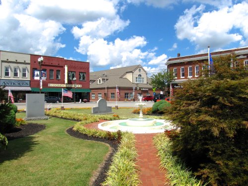 A small park with a fountain surrounded by greenery, with shops and blue skies in the background.
