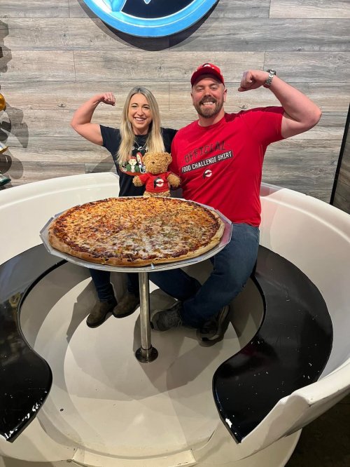 A man and woman flex their muscles beside a large pizza on a round table, smiling and posing with a teddy bear.