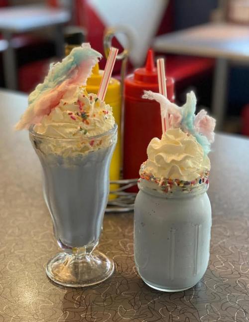 Two colorful milkshakes topped with whipped cream, sprinkles, and cotton candy, set against a retro diner backdrop.