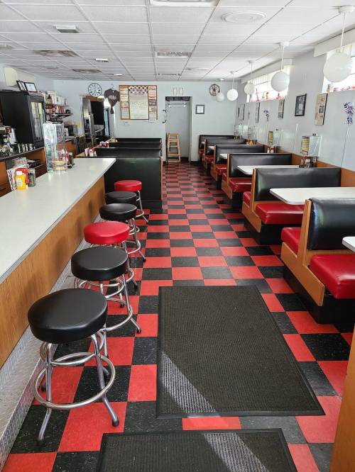 A retro diner interior with red and black checkered floors, bar stools, and booth seating along the walls.