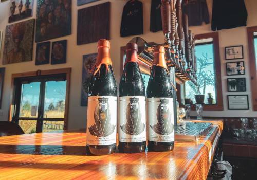 Three bottles of dark beer with artistic labels sit on a wooden bar, surrounded by brewery decor and taps in the background.
