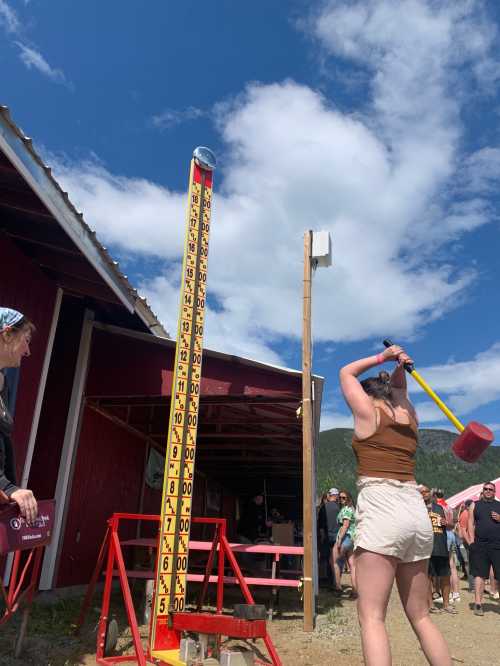 A person swings a mallet at a height measurement game outside a red barn, with a crowd and blue sky in the background.