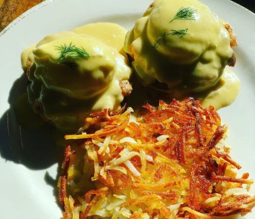 Two eggs Benedict topped with hollandaise sauce and a side of crispy golden hash browns on a white plate.
