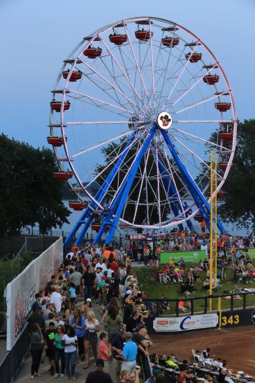 A crowded fairground scene featuring a brightly lit Ferris wheel against a twilight sky.