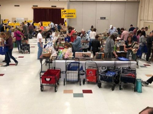 A busy book fair with people browsing tables filled with books and carts in a large indoor space.