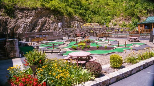 A mini-golf course surrounded by flowers and rocky terrain, featuring various putting greens and seating areas.