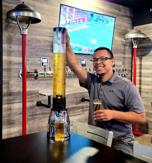 A man smiles while holding a glass of beer next to a tall beer tower in a bar with a TV showing a sports game.