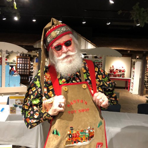 A festive person with a white beard, sunglasses, and a colorful outfit stands in front of a holiday-themed display.