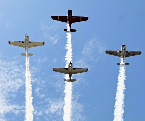 Four vintage aircraft flying in formation, trailing white smoke against a blue sky with scattered clouds.