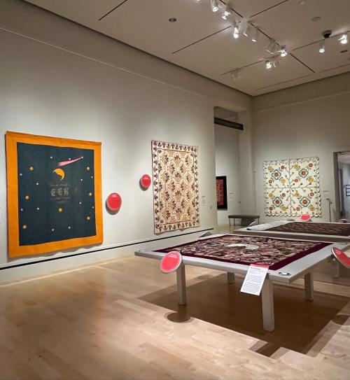 A gallery with colorful textile artworks displayed on the walls and a table featuring more textiles in the center.