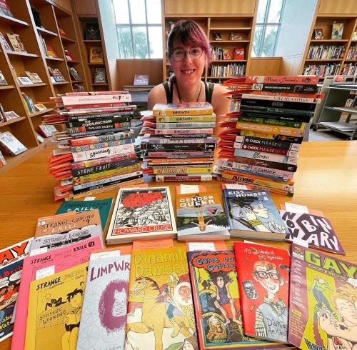 A person with pink hair smiles behind a table stacked with colorful graphic novels and comics in a library setting.