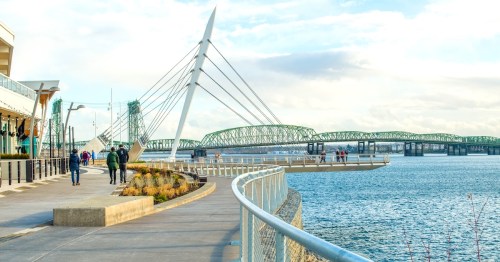 A scenic waterfront view featuring a bridge, walking path, and people enjoying the area under a cloudy sky.