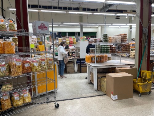 A grocery store interior with shelves of snacks and customers browsing. Boxes and a cleaning cart are visible.