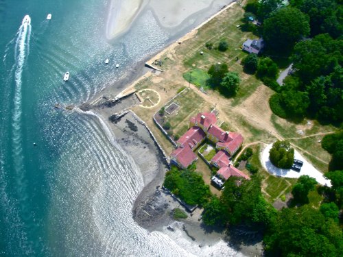 Aerial view of a coastal property with a red-roofed house, sandy shore, and boats in the water nearby.