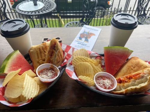 Two baskets of food with sandwiches, chips, watermelon slices, and dipping sauce, alongside two coffee cups.
