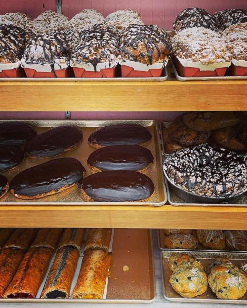 A display of assorted baked goods, including chocolate-covered pastries, muffins, and sweet rolls on wooden shelves.