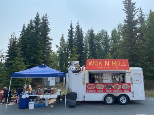 A food truck named "Wok N Roll" serving Chinese cuisine, with a blue tent and tables set up nearby in a forested area.