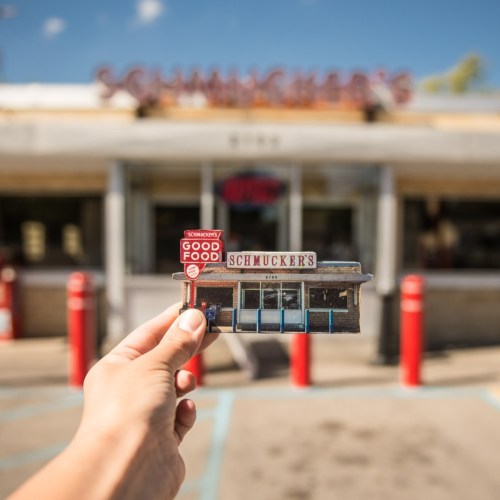 A hand holds a postcard of Schmucker's restaurant in front of the actual building, with a clear blue sky above.