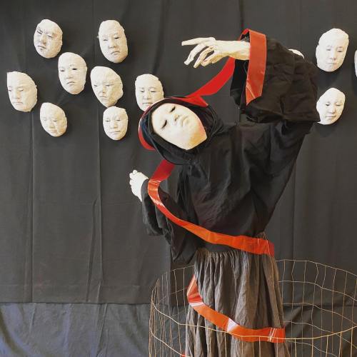 A figure in a black cloak poses dramatically against a dark backdrop with several white, expressionless masks.