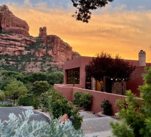 A scenic view of a modern building surrounded by trees, with red rock formations and a colorful sunset in the background.