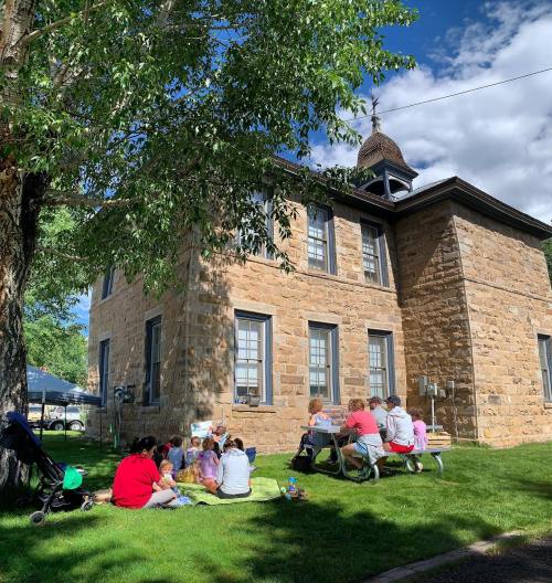 A group of people sits on the grass near a stone building, enjoying a sunny day under a tree.