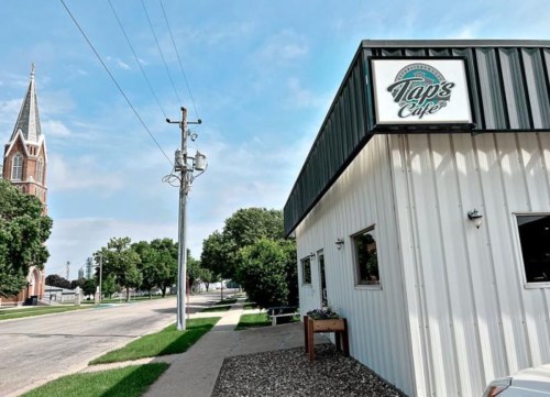 Exterior view of Taps Cafe with a church steeple in the background, set along a tree-lined street.