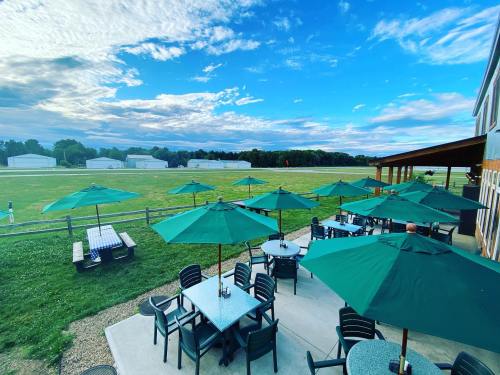 Outdoor seating area with green umbrellas, tables, and a grassy field under a blue sky with scattered clouds.