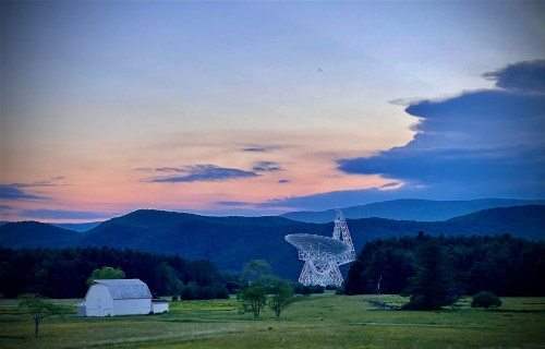 A white radio telescope stands in a green field, surrounded by mountains under a colorful sunset sky.