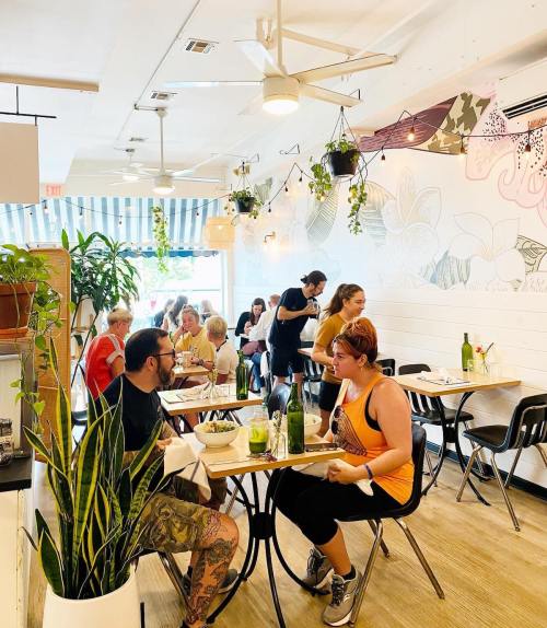A bright, modern café with diners at tables, plants, and a server attending to customers.