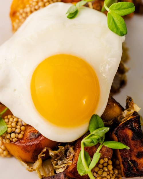 A close-up of a poached egg on roasted vegetables, garnished with fresh greens and mustard seeds.