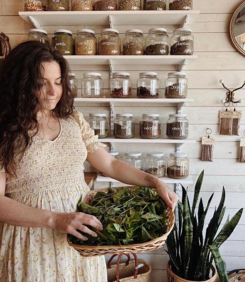 A woman in a floral dress holds a basket of greenery in a shop with jars of herbs and plants on the wall.