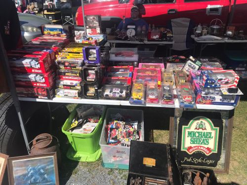 A table filled with various toy cars and collectibles at a flea market, with a vendor in the background.