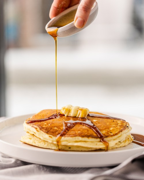 A stack of pancakes topped with a pat of butter, drizzled with syrup from a small bowl.