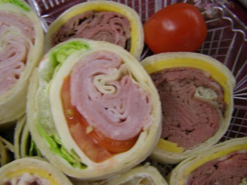 Close-up of rolled sandwiches with layers of ham, roast beef, lettuce, and tomato, with a whole tomato beside them.