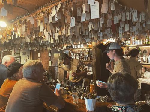 A busy bar scene with patrons chatting and a bartender serving drinks, surrounded by hanging tags on the walls.