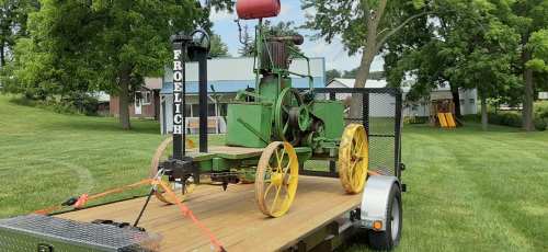 A vintage green farming machine on a trailer, set on a grassy area with trees and buildings in the background.
