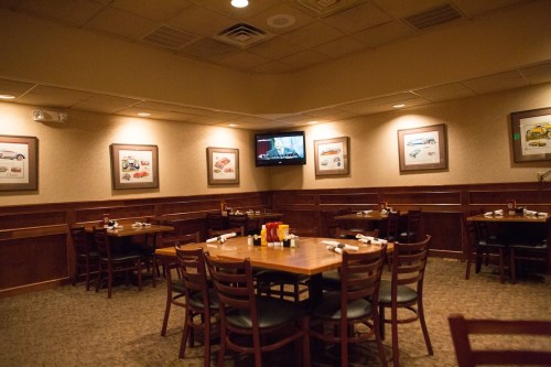 A cozy restaurant interior with wooden tables, chairs, and framed car art on the walls. A TV is mounted in the corner.