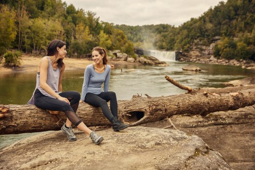 Two women sit on a log by a river, smiling and enjoying nature, with a waterfall and trees in the background.