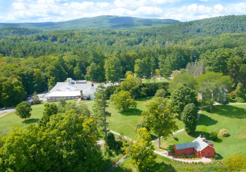 Aerial view of a lush green landscape with a large building and a red barn surrounded by trees and rolling hills.