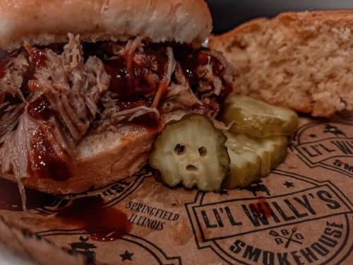 Close-up of a pulled pork sandwich with barbecue sauce, pickles, and cornbread on a branded paper plate.