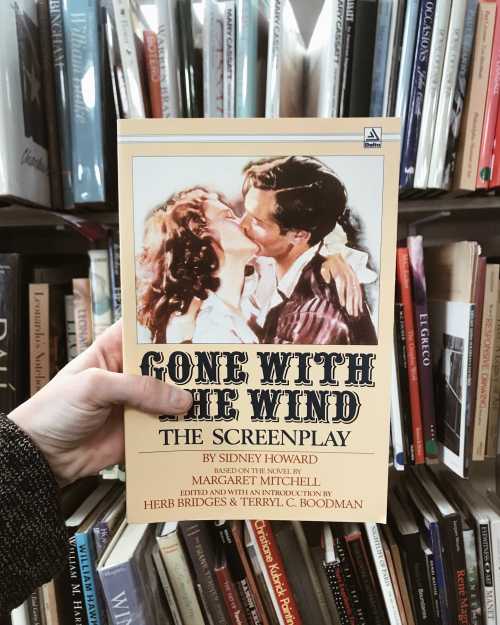 A person holds a copy of "Gone with the Wind: The Screenplay" in front of a bookshelf filled with books.