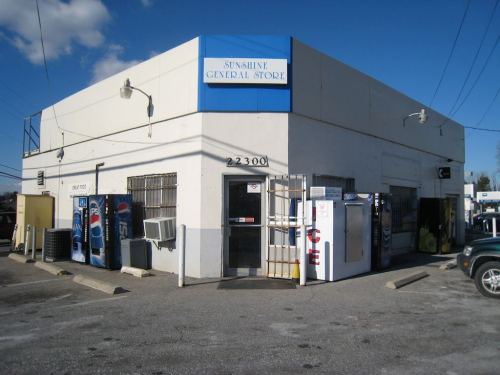 A corner store with a blue sign reading "Sunshine General Store," featuring vending machines and a parking area.