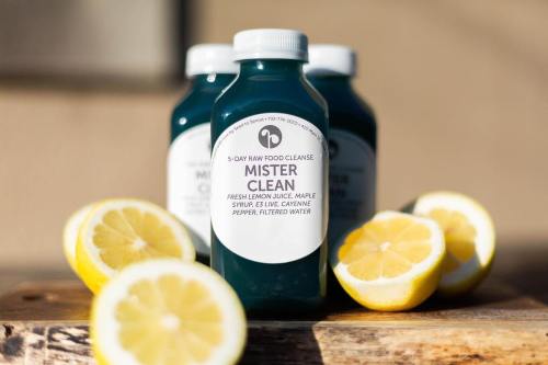 Three bottles of "Mister Clean" juice surrounded by fresh lemon halves on a wooden surface.