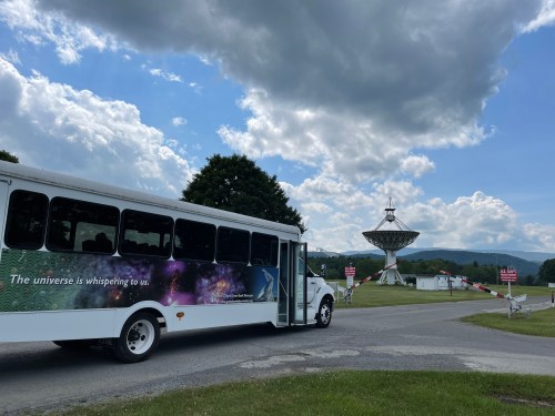 A bus with a cosmic-themed design parked near a large satellite dish under a cloudy sky.